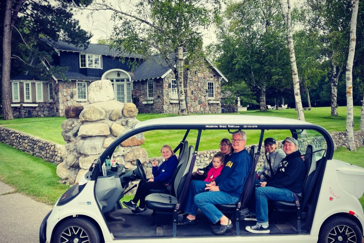 Five people sitting in a parked golf cart on a driveway near a stone house and trees.