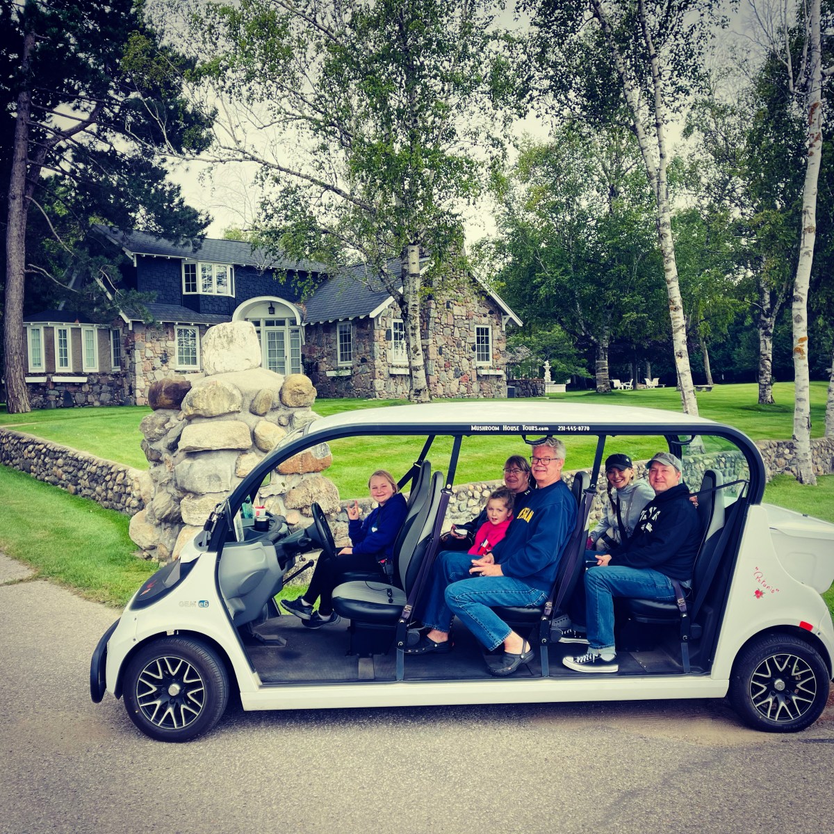 Five people sitting in a parked golf cart on a driveway near a stone house and trees.