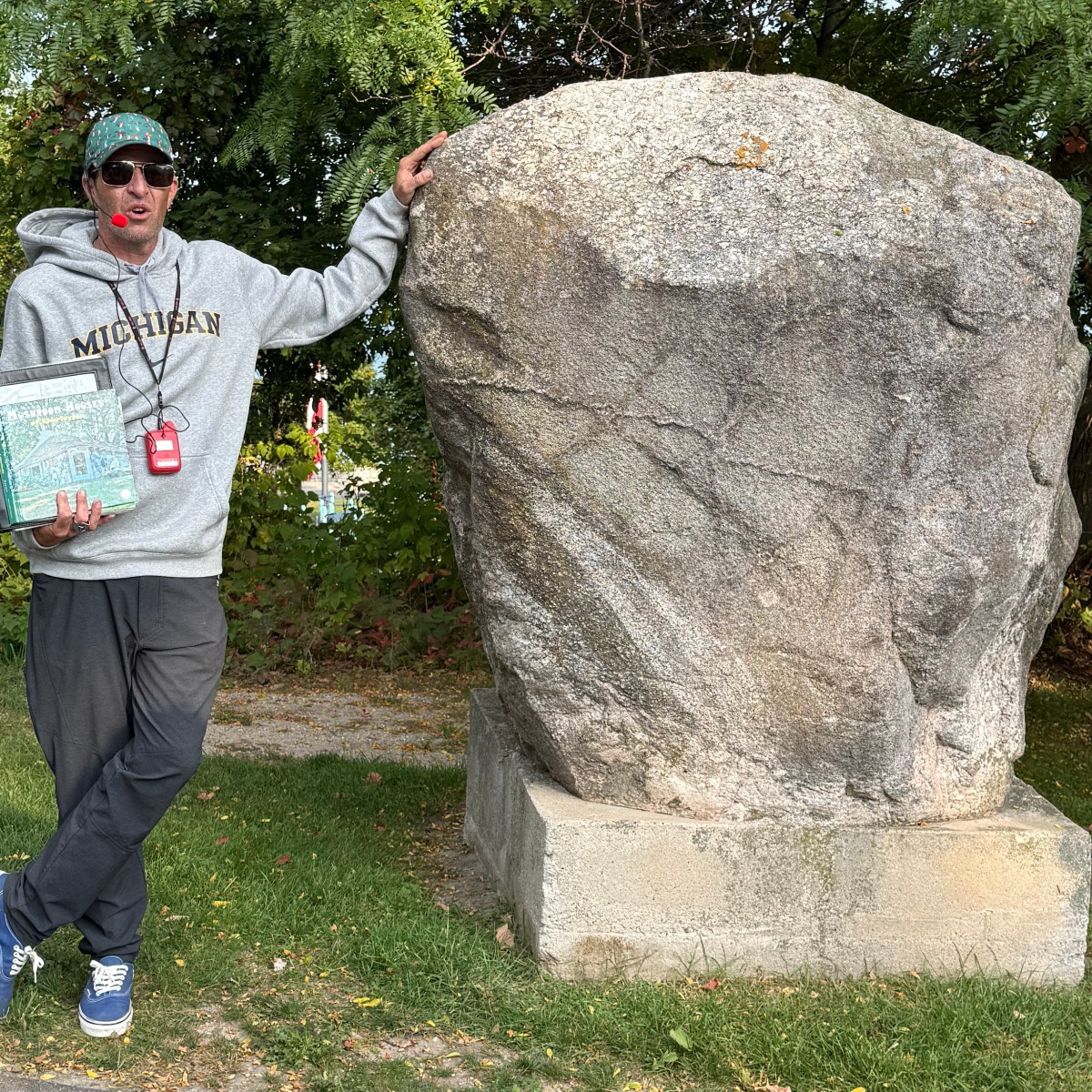 Person in Michigan hoodie leans on a large rock outdoors, holding a binder with a whistle around their neck.