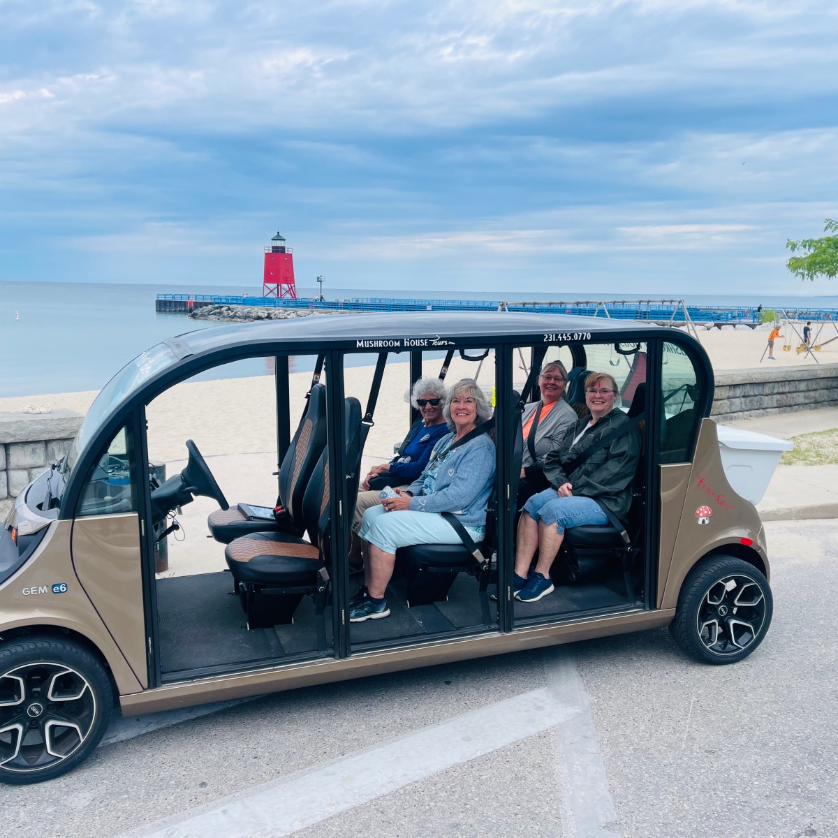 Four people seated in an open-air vehicle near a beach with a red lighthouse in the background.