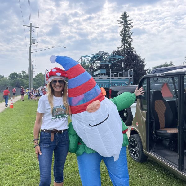 Woman in mushroom hat poses with inflatable gnome beside a small car on a grassy field.