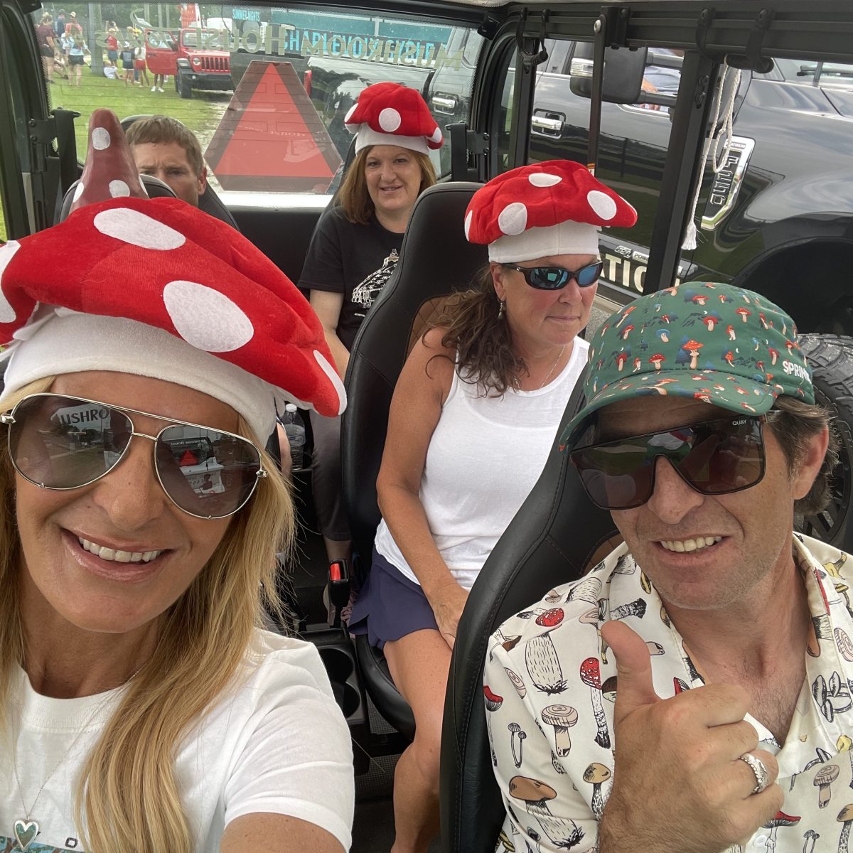 Group of people in a vehicle wearing mushroom-themed hats and shirts, smiling at the camera.