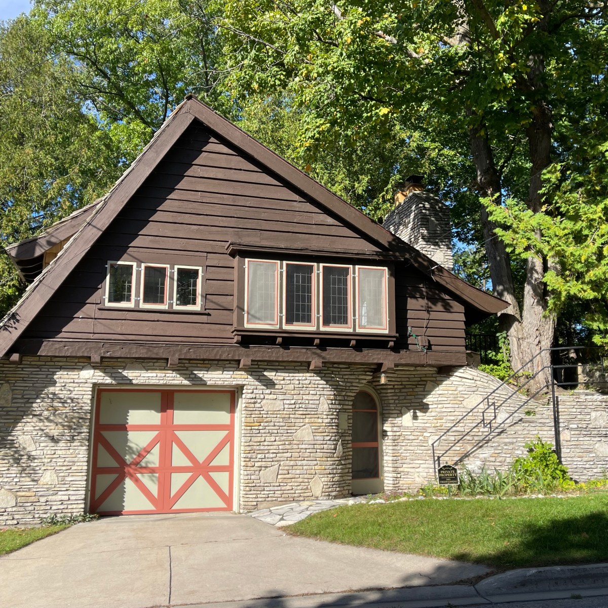 Brown and stone house with garage and sloped roof, surrounded by trees.