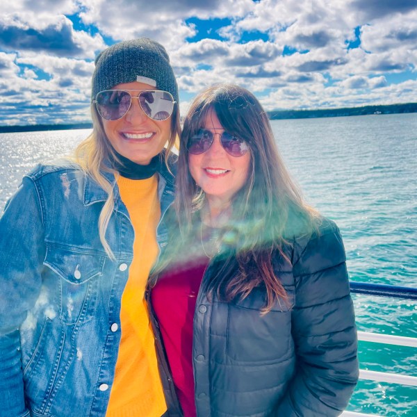 Two women in sunglasses standing on a boat with a sunny sky and water in the background.