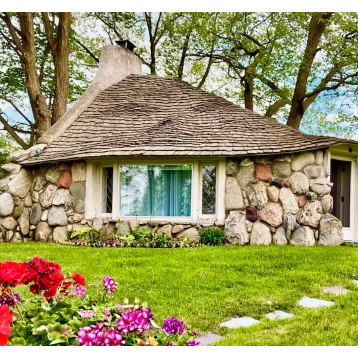 Stone cottage with shingled roof, surrounded by trees and colorful flowers.