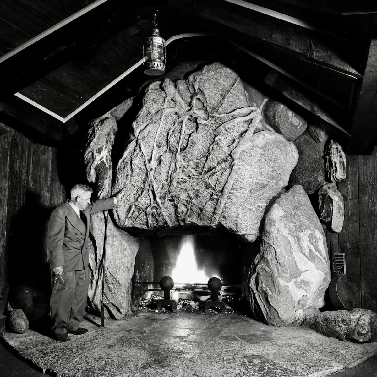 Man standing by large stone fireplace with burning fire, surrounded by rocks.
